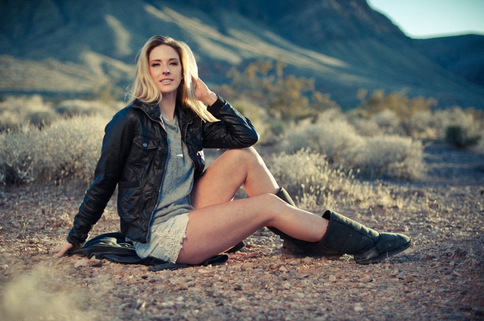 Stunning blonde model sitting on desert ground with sun behind her wearing leather jacket