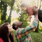 Father holds son in the air as mother smiles in green yard for family portrait in Nashville Tennessee